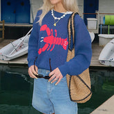 Person wearing a blue sweater with a red lobster design, holding sunglasses and a straw bag, standing by a dock with boats.